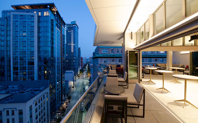 A modern cityscape at dusk with high-rise buildings and a spacious balcony featuring seating and tables, overlooking the illuminated street below.