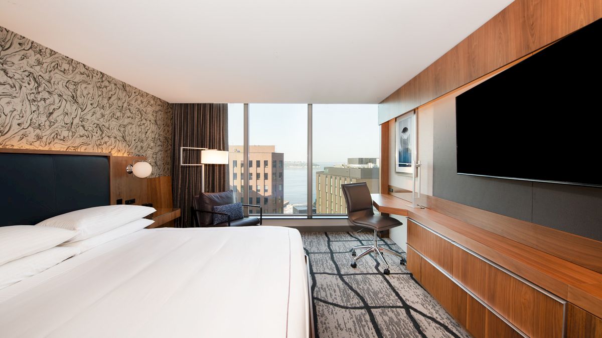 A modern hotel room features a large bed, desk, chair, flat-screen TV, and a window with a city view. Light and wood accents complete the decor.