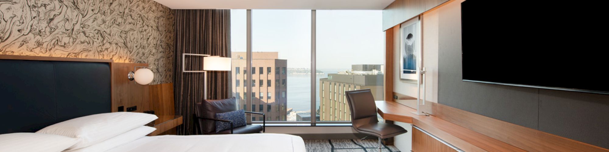 A modern hotel room features a large bed, desk, chair, flat-screen TV, and a window with a city view. Light and wood accents complete the decor.